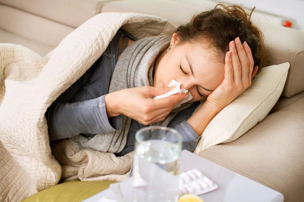 Woman resting on a couch during cold and flu season with tissues, water, and medication nearby, representing fatigue, congestion, and winter illness symptoms.