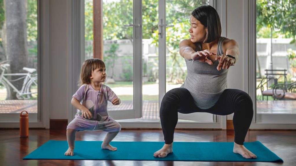 Pregnant woman performing a deep squat exercise on a yoga mat at home while a young child imitates her movement beside her. The image highlights the importance of staying active, maintaining mobility, and preparing the body for pregnancy, birth, and motherhood through movement.