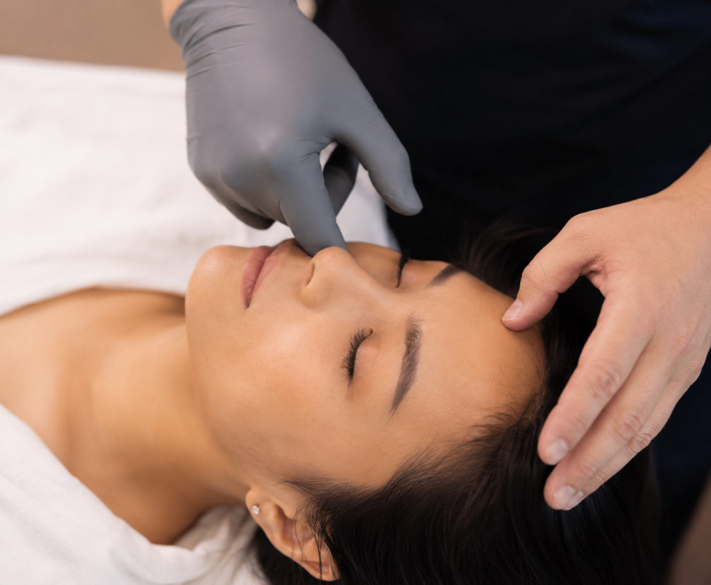 Practitioner performing a gentle TMJ and cranial assessment on a woman lying comfortably on a treatment table, with one hand supporting the head and the other assessing facial structures.