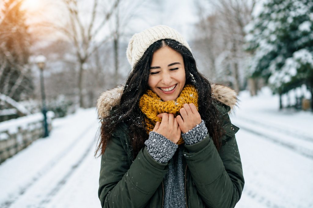 Smiling woman wearing a green winter coat, knitted beanie and mustard scarf, standing outdoors on a snowy winter morning while enjoying fresh air and gentle snowfall.